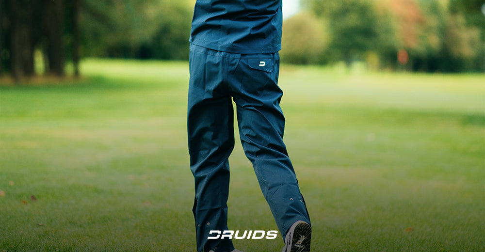 Close-up view of a golfer wearing navy golf pants with a pocket detail showcasing the DRUIDS logo on a green golf course background.